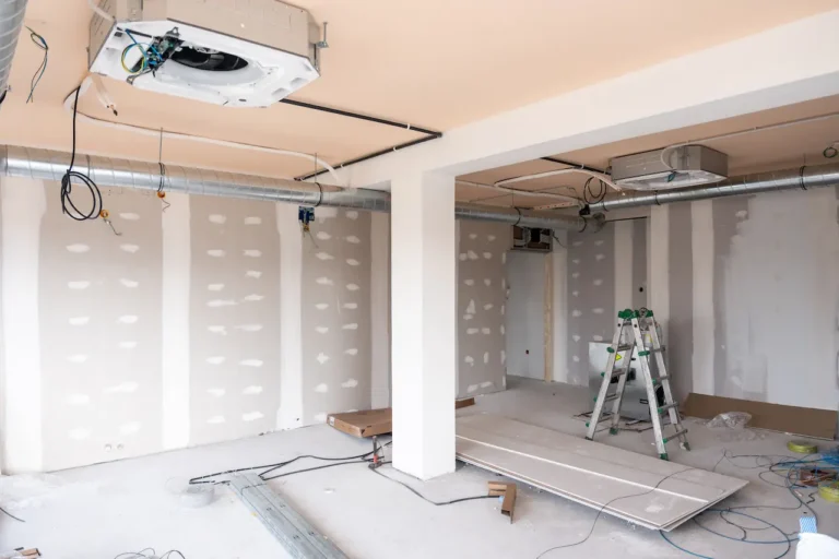 Under-construction interior with drywall panels, exposed wiring, and a ladder in the center of the room, unfinished ceiling.